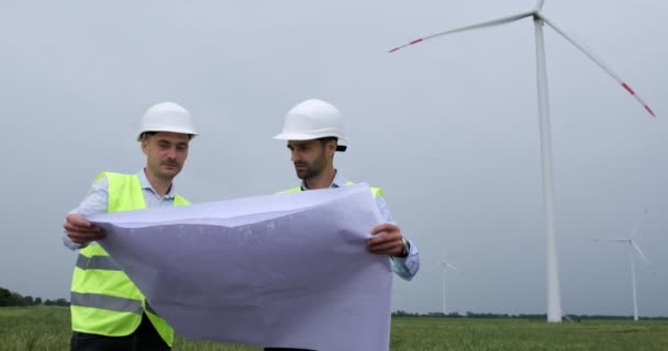Muži v bílých helmách drží projektový list a diskutují o větrném mlýně — Stock video