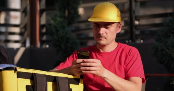 Mensajero con el teléfono apoya el brazo inclinado en la termobolsa en la cafetería — Vídeos de Stock