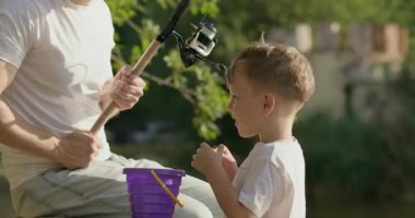 Anaokulu çocuğu balık tutma gezisi sırasında babasına küçük plastik oyuncak balıklar veriyor. Baba ve küçük oğlu sakin gölün kıyısındaki ahşap iskelede oturur ve balık avlama gününün tadını çıkarırlar.