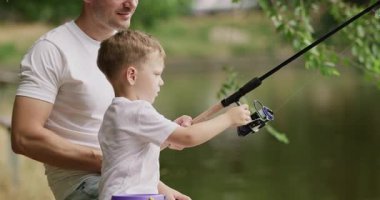 Baba, saydam suya işaret ediyor. Oğul balıkların suyun altında yavaşça yüzdüğünü gösteriyor. Bakıcı baba küçük çocuğun elini oltaya takar ve oltaya tutunmasına yardım eder.