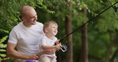 Mutlu baba, nehir kıyısında balık tutarken oğlunu kollarına aldı. Önemseyen baba, anaokulu öğrencisinin oltayla boğuşmasını ve heyecanla gülmesini izliyor.