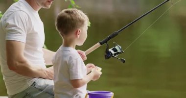 Anaokulu çocuğu nehir kıyısındaki ahşap iskelede oturan oyuncak balıkla oynadığı gibi mutlu bir şekilde gülüyor. Babam balıkçı oltasıyla sakince iskelede oturur ve güneşli bir günün tadını çıkarır.