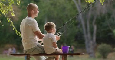 Baba ve küçük çocuk ahşap bankta su oltasının yanında oturuyorlar. Sevimli baba ve oğul nehir kıyısındaki bankta huzurlu bir balık tutma anının tadını çıkarıyorlar.