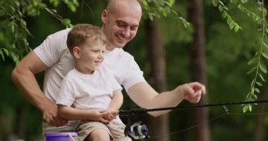 Baba olta ipinin ucundaki balık kıvrımına doğru gülümsüyor. Babam gülüyor ve oğlanı oltayı çekmeye teşvik ediyor. Çocuk büyük gözlerle hareket eden balığa bakıyor.