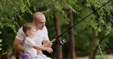 Cesur bir adam oltayı elinde tutuyor ve çocuk merak ve heyecanla suyu seyrediyor. Babam ve küçük oğlum nehir kıyısında oturuyorlar. Etrafı uzun yeşil ağaçlarla çevrili.