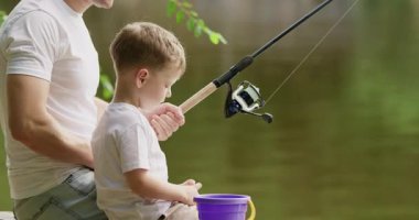 Anaokulu çocuğu gülümser ve renkli balık şeklinde küçük bir oyuncakla oynar. Küçük çocuk nehir kıyısında balık avlarken babasının yanındaki eski ahşap iskelede oturur.