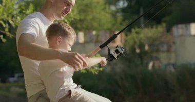 Baba ve oğul ahşap iskelede birlikte otururken gülümsüyorlar. Ebeveyn, sevimli çocuğa gölün ışıl ışıl yüzeyinden küçük balıkları çıkarmasında yardım eder.