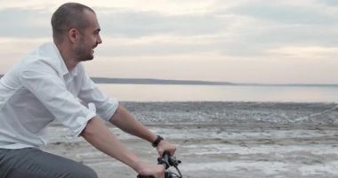 Gülümseyen ofis çalışanı sıcak güneş ışığı altında yumuşak kumlu sahil boyunca neşeyle bisiklet sürüyor. Mutlu adam, yumuşak hareketler sırasında huzurlu kıyı esintisini kucaklıyor.