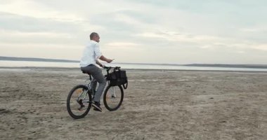 Odaklanmış adam bisiklet sürüyor parlak kumlu sahil şeridi boyunca dengeleyici çanta gidonu üzerinde. Kendine güveni olan bir adam cebinden telefonu kapar ve hemen cevap verir.