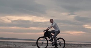 Serene ofis çalışanı, altın gece parıltısıyla aydınlatılan kıyı şeridinin yakınında sessizce bisiklet sürüyor. Acelesi olmayan adam, gün batımında sakin sahil yolunu kucaklıyor.