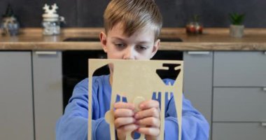 Odaklanmış çocuk evdeki kontrplak kağıdının uçak modelinin yuvarlak kısmını çiziyor. Birinci sınıf öğrencisi mutfak masasında oturup eski model oyuncak uçak monte etmeyi öğreniyor.