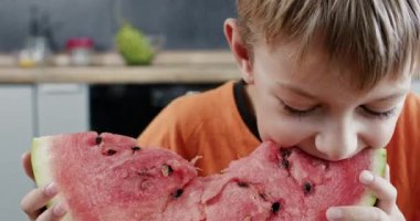 Tişörtlü çocuk siyah tohum arayan karpuz diliminin tadını alıyor. Çocuk, bir sonraki ısırıktan önce çekirdek ceplerini incelemek için kırmızı hamura dil basar.