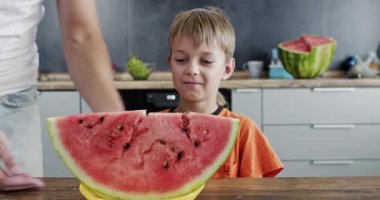 Kadın mutfak masasına kocaman bir karpuz takozu koyuyor ve çocuk aç bir şekilde gülümsüyor. Çocuk elleriyle kabuğu kavrayıp büyük ısırıklarla karpuz dilimi yiyor.