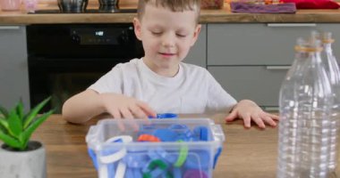 Genç çocuk ahşap masadaki plastik şişelerin yanındaki açık kutuya mavi başlık bırakıyor. Anaokulu çocuğu, geri dönüşüm için küçük kapağı kabın içine yerleştirme işlemini tamamladı