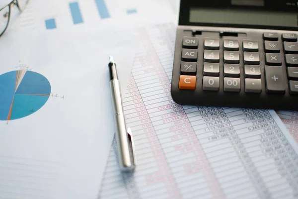 Finansal muhasebe, yakın çekim hesap makinesi, kağıt üzerinde sayılar, birçok sayı, grafik, kalem, kağıt üzerinde gözlük, ofisteki masaya yerleştirilen belgeler, bulanık arkaplan.