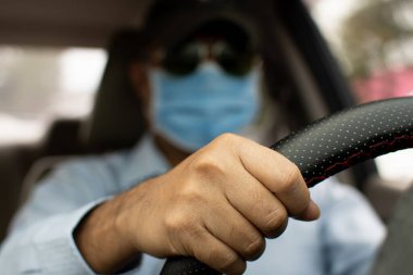 Trafik sıkışıkken arabayı dikkatle kontrol etmek için direksiyonu tutmak için iki elini kullanan bir adam gündüz vakti. Yüz maskesi takan bir erkek. Yaklaşın ve elinize odaklanmayı seçin. Bulanık arkaplan