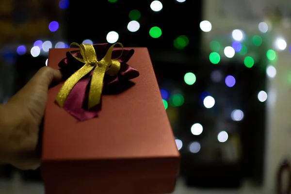 The hand of man holding red color Presents celebrates box with red and gold ribbons on Christmas day, take close-up photography, bokeh lights, and blurred background