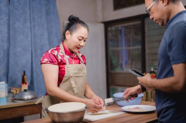 Asyalı bir çift ev mutfaklarında taze ve sağlıklı bir yemek hazırlıyor. Kadın yemek keserken, adam akıllı telefonu yemek tarifi olarak kullanıyor. Ev hayatının tadını çıkarıyor..