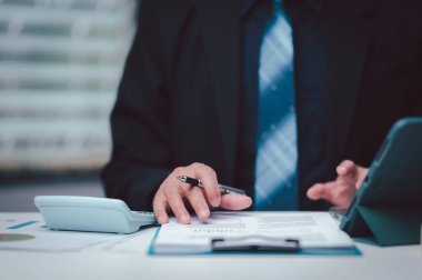 Profesyonel bir işadamı masasında hesap makinesi ve dijital tablet kullanarak kurumsal iş stratejisinin doğruluğunu ve planlamasını sağlayan bir mali raporu analiz ediyor..