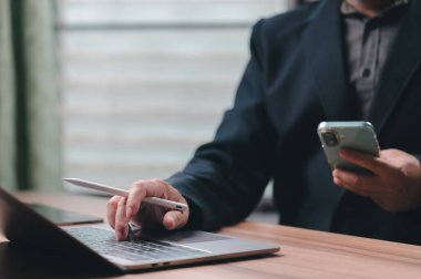 Koyu takım elbiseli profesyonel iş adamı ahşap masadan çalışır. Eller dizüstü bilgisayarda klavye yazacak, akıllı telefon tutacak, stil kalemi kavrayacak. Modern işyeri teknolojisi uzak işleri kolaylaştırıyor