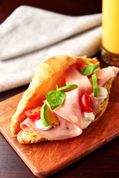 Appetizing croissant with mortadella sausage, cream cheese, avocado, tomato and fresh basil leaves served with glass of orange juice. Tasty breakfast.