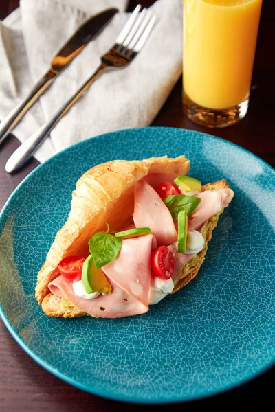 Appetizing croissant with mortadella sausage, cream cheese, avocado, tomato and fresh basil leaves served with glass of orange juice. Tasty breakfast.