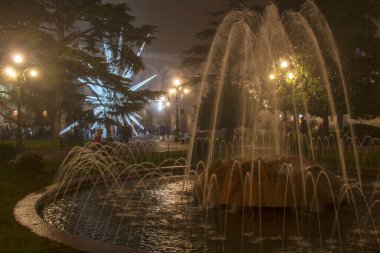 Parlayan lambalar, bayram ışıkları ve sisli, atmosferik bir meydanda akşam yürüyüşünün keyfini çıkaran insanların siluetleriyle çevrili bir şehir parkında gece vakti aydınlatılmış bir çeşme su sıçratır..