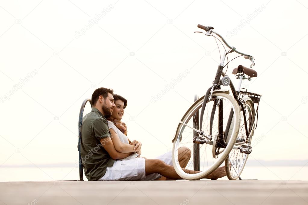 Couple in love sitting on a boardwalk with a bike at the lake