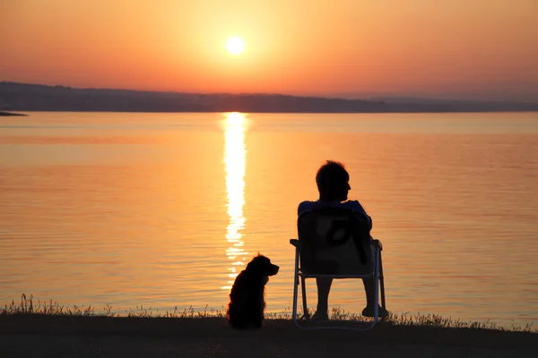 El hombre y su amigo perro admiran el amanecer en el mar — Foto de Stock