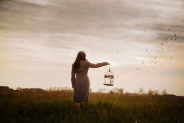 Kız gün batımında boş bir kafesle sonsuzluğa doğru yürürken gökyüzündeki özgür kuşları seyrediyor..