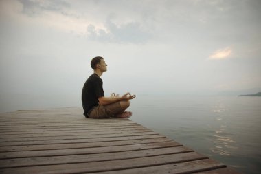 Genç adam deniz kıyısında meditasyon yapıyor, sakin bir şekilde nefes alıyor ve bağ kuruyor.