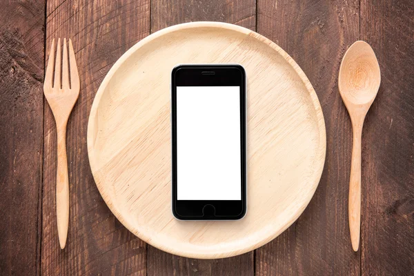 Еда смартфона на комплекте вилки, ложки и блюдца древесины на деревянной т — стоковое фото