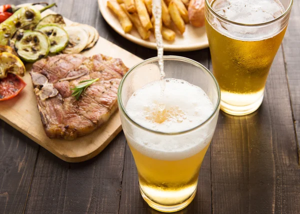 Cerveja sendo derramada em vidro com bife gourmet e batatas fritas — Fotografia de Stock