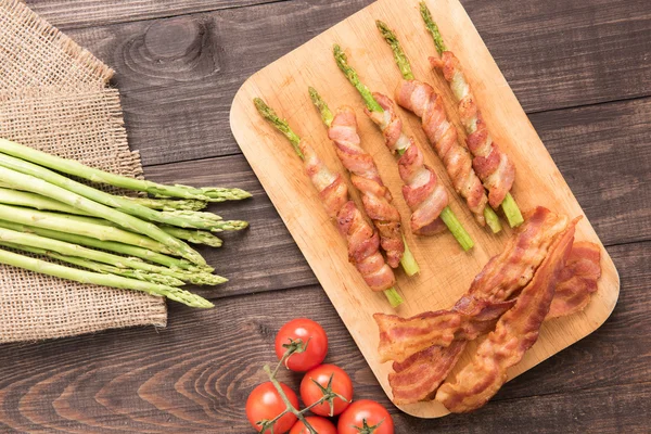 Asparagus wrapped in bacon with tomato on wooden background — Stock Photo, Image