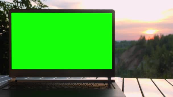 Portátil negro con una pantalla verde está en una mesa plegable al atardecer en la cima de la montaña. — Vídeos de Stock