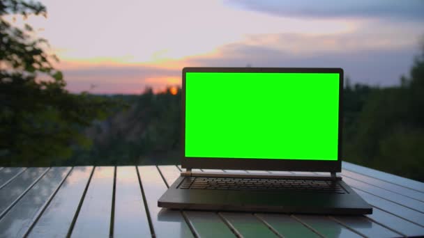 Portátil negro con una pantalla verde está en una mesa plegable al atardecer en la cima de la montaña. — Vídeos de Stock