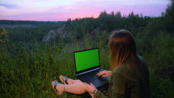 Chica joven trabaja en un ordenador portátil con una pantalla verde en la cima de una montaña. — Vídeos de Stock