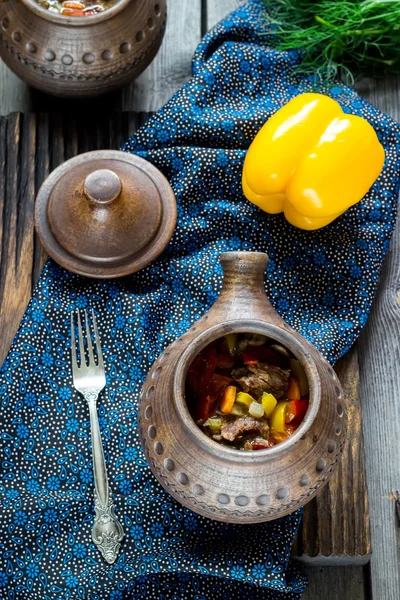 Olla de cerámica con carne y verduras, pimiento amarillo y servilleta en mesa de madera oscura . — Foto de Stock