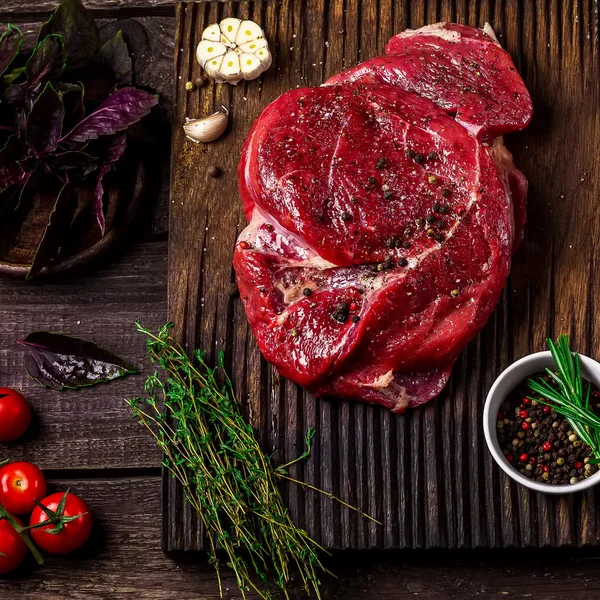 La carne de vaca en la tabla de cortar, los tomates, las hierbas y las especias en la mesa de madera oscura. Estilo rústico . — Foto de Stock