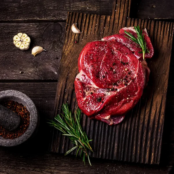 Βόειο κρέας σε μια σανίδα κοπής, ντομάτες, βότανα, σκόρδο και μπαχαρικά σε σκούρο ξύλινο τραπέζι . — Φωτογραφία Αρχείου