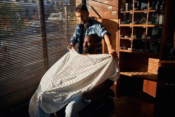 Skäggig frisör sätter frisörmantel på klienten i frisörsalongen — Stockfoto