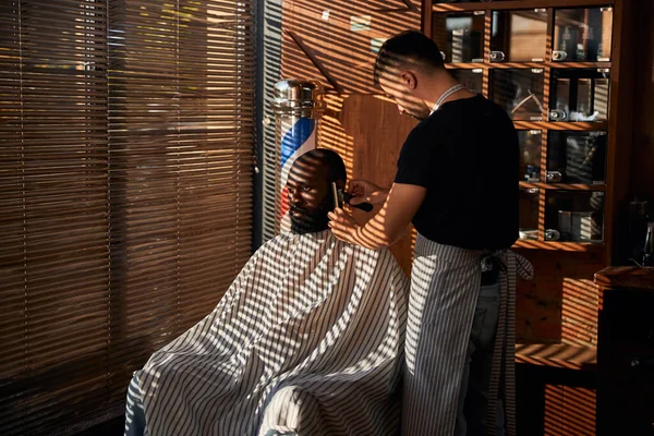 Man kapper in schort trimmen klant baard in kapperszaak — Stockfoto