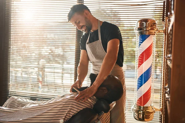 Cheerful barber trimming client beard in barbershop — Stock Photo, Image