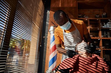 Tıbbi maskeli berber berber berber dükkanında müşterinin sakalını tıraş ediyor.