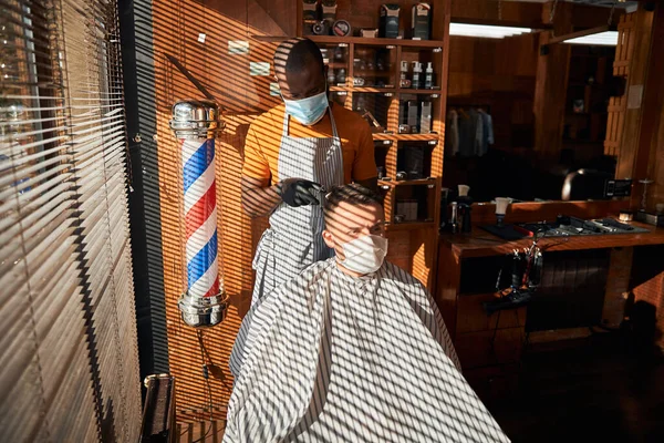 Barbeiro em máscara médica corte de cabelo do cliente na barbearia — Fotografia de Stock