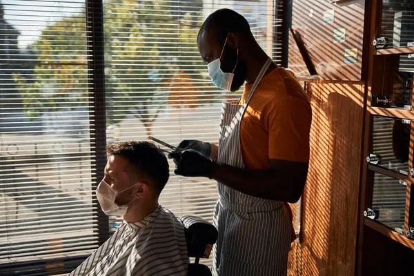 Homem jovem barbudo em máscara médica recebendo corte de cabelo na barbearia — Fotografia de Stock