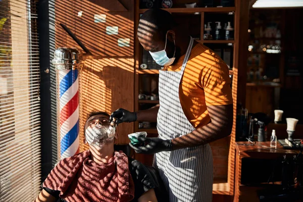 Mann-frisør i medisinsk maske som bruker barberskum på menneskets ansikt – stockfoto