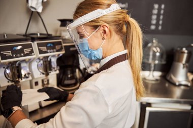 Beyaz gömlekli ve önlüklü güzel bir kadın otomatik kahve makinesinin yanında duruyor.