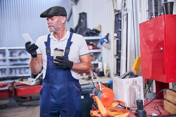 Baard monteur met behulp van mobiele telefoon in auto reparatiewerkplaats — Stockfoto