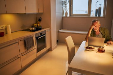 Yorgun düşmüş yaşlı vatandaş dizüstü bilgisayarla çalışırken baş ağrısı çekiyor.